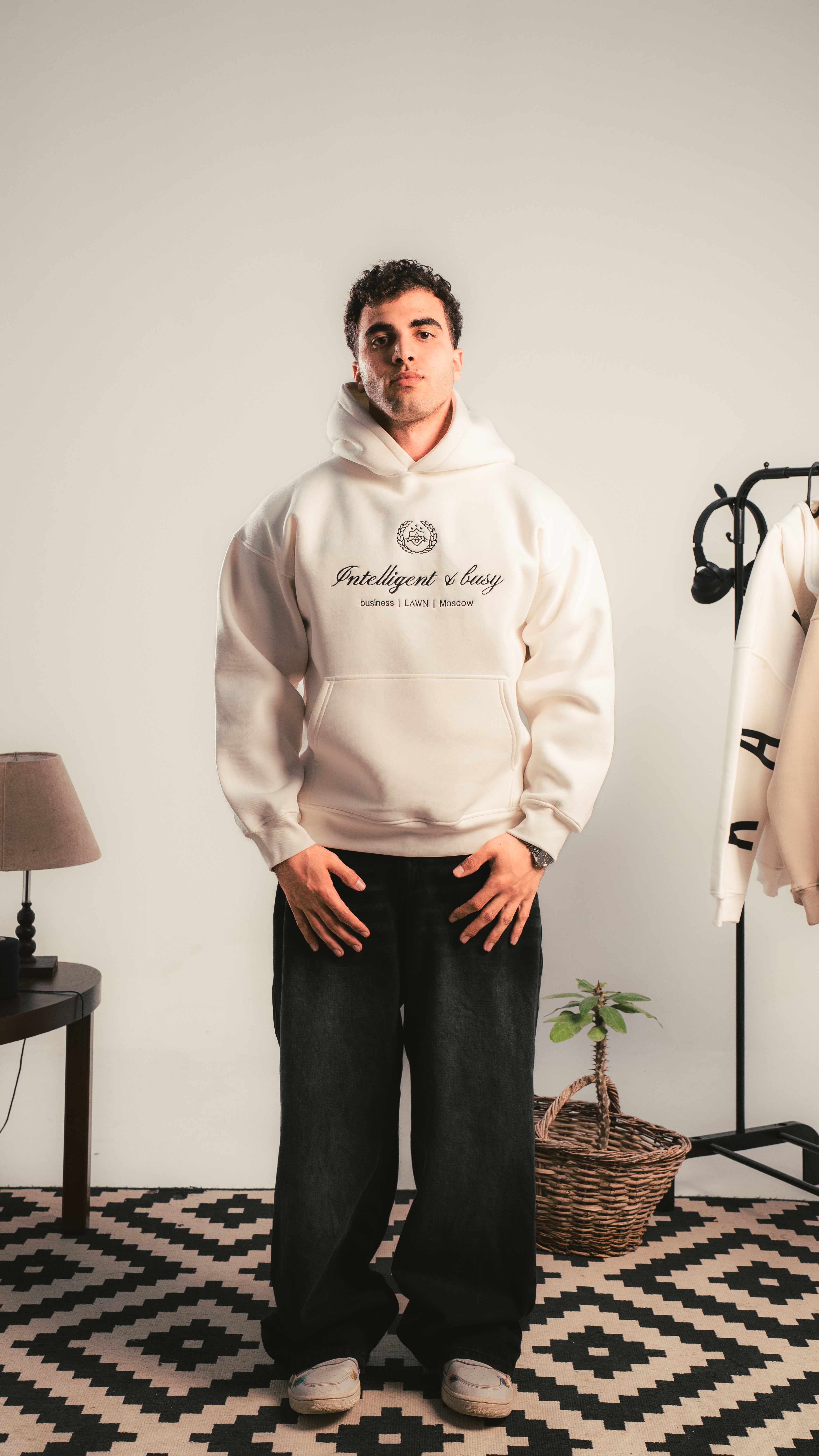 Person wearing a white hoodie with text, standing in a room with a lamp and plant.