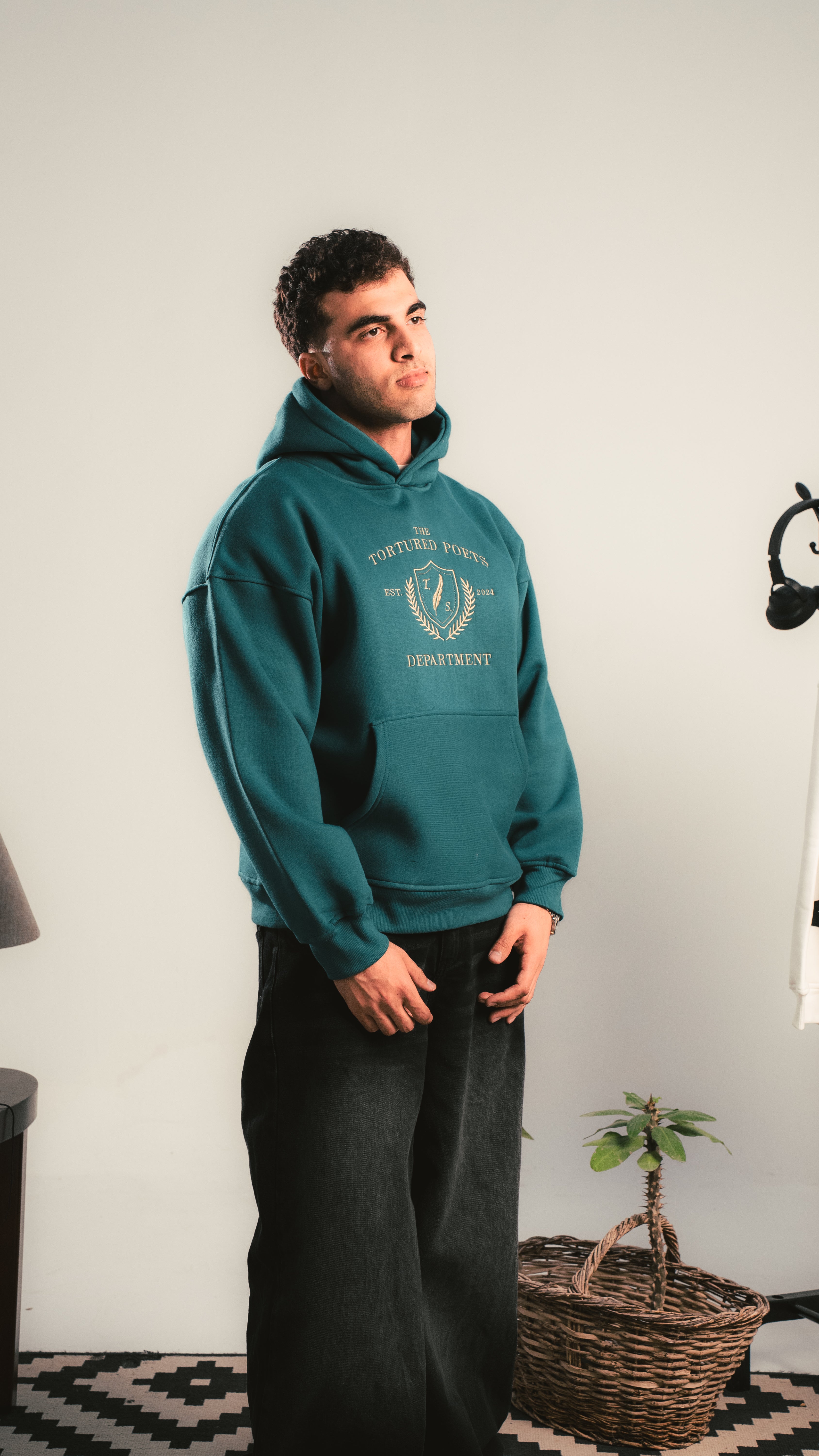 Person wearing a teal hoodie with a logo, standing indoors against a white wall.