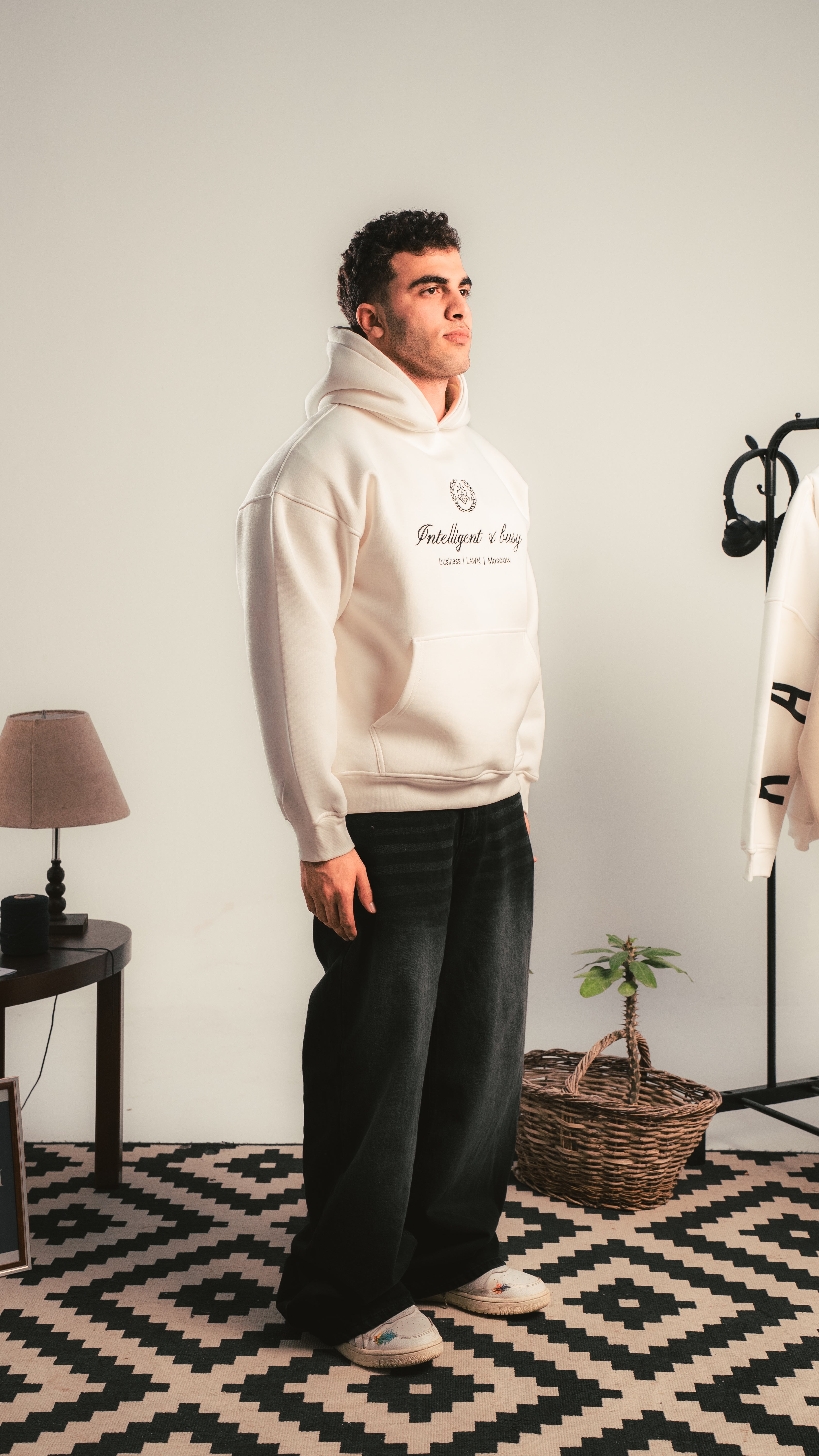 Person wearing a white hoodie with text in a room with a lamp, plant, and basket.