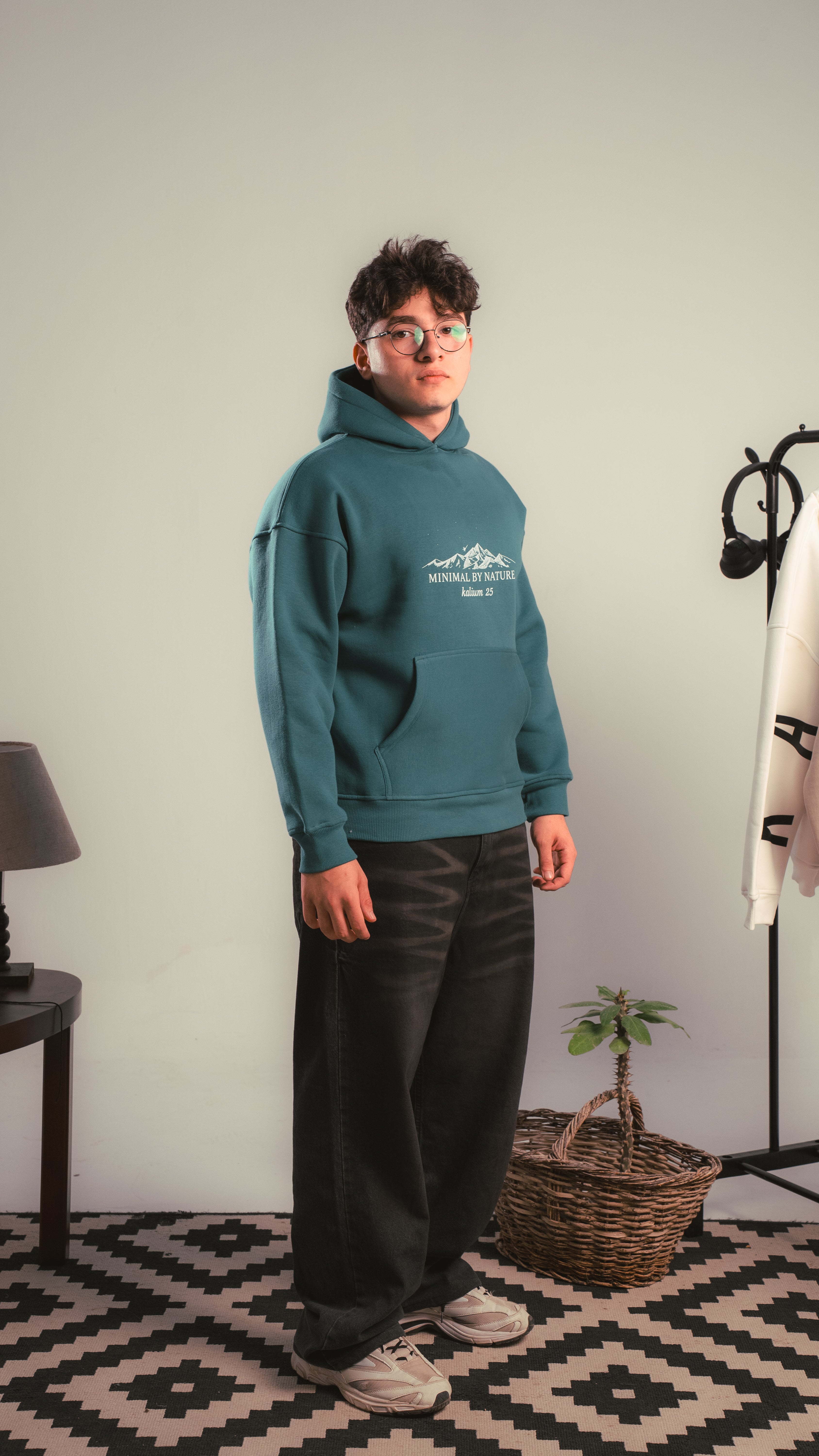 Person wearing a teal hoodie with a logo in a room with a lamp, basket, and plant.