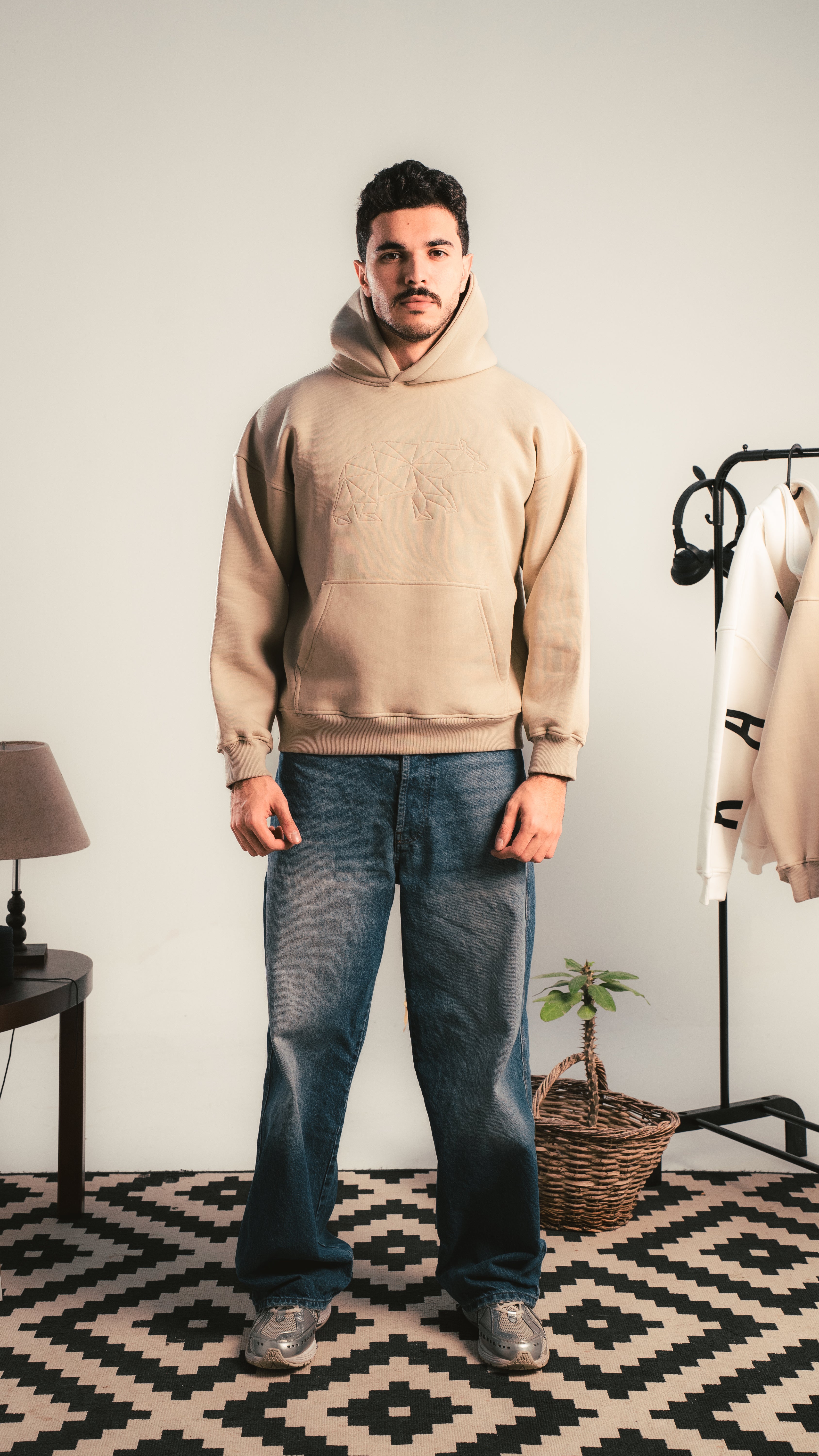 Person wearing a beige hoodie with an embossed teddy bear graphic, standing indoors in a minimal room with a lamp and clothing rack