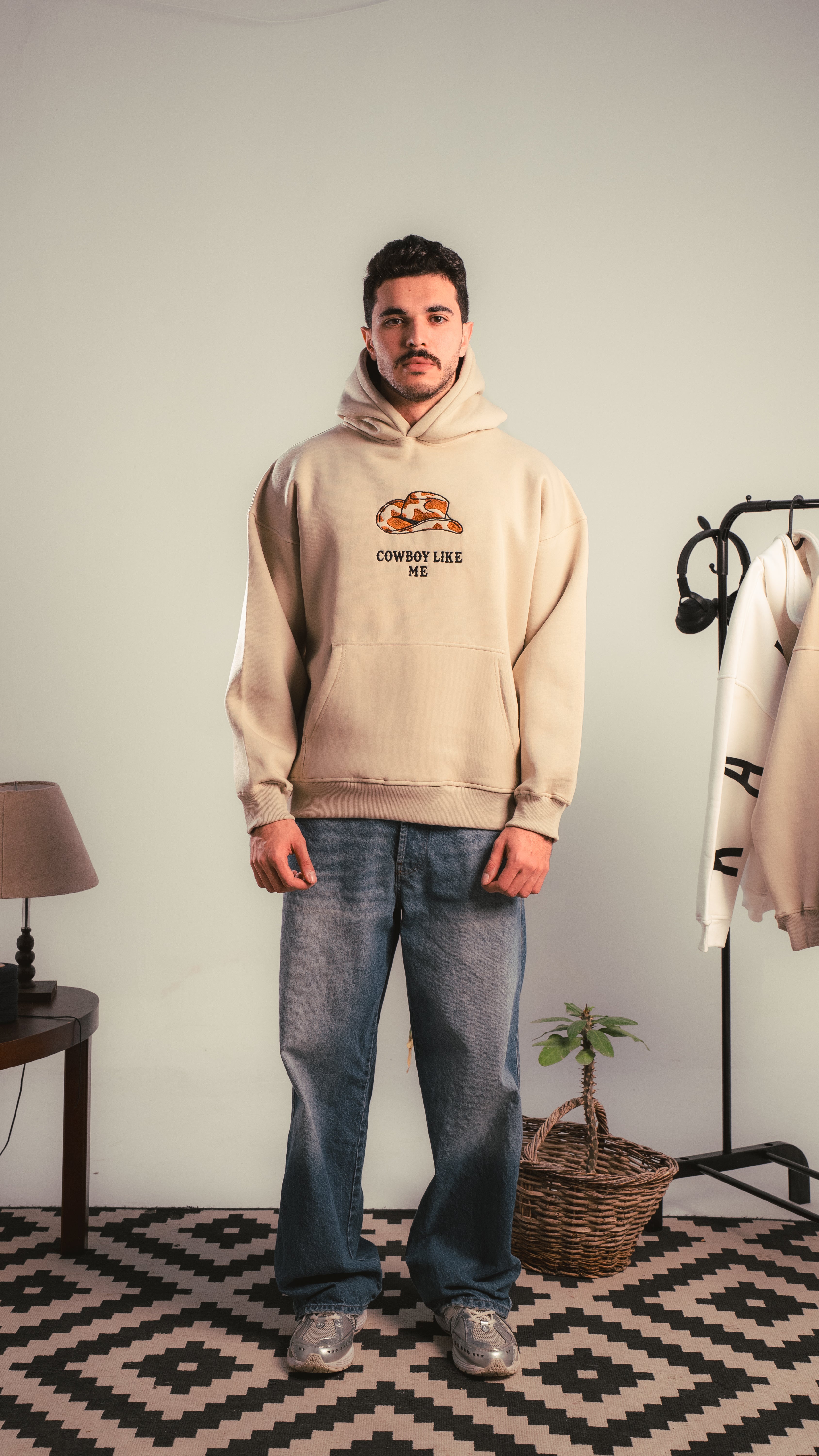 Person wearing a beige hoodie with ‘Cowboy Like Me’ print, standing indoors