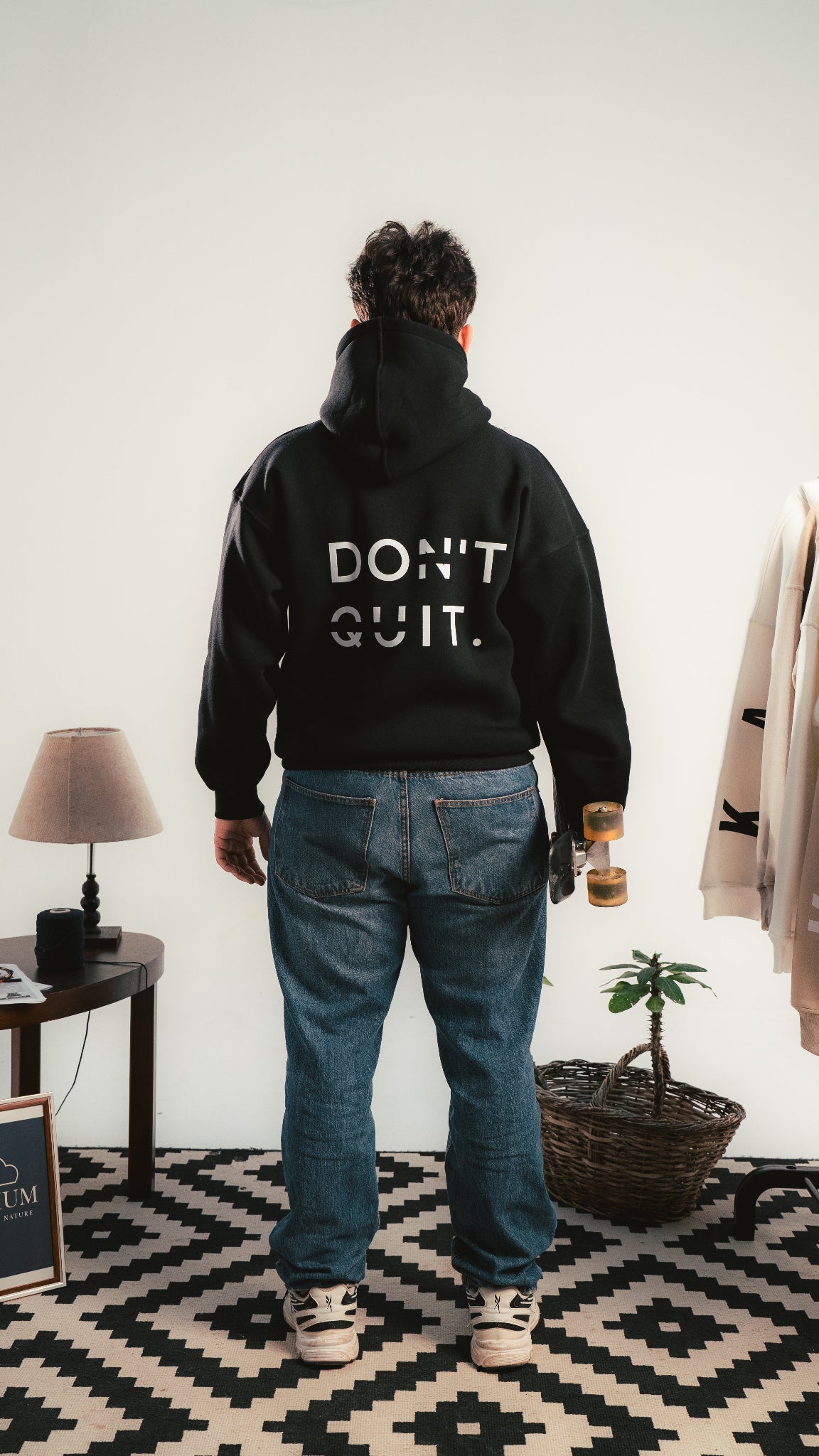 Person wearing a black hoodie with 'DON'T QUIT.' text, standing in a room with a lamp and plant.
