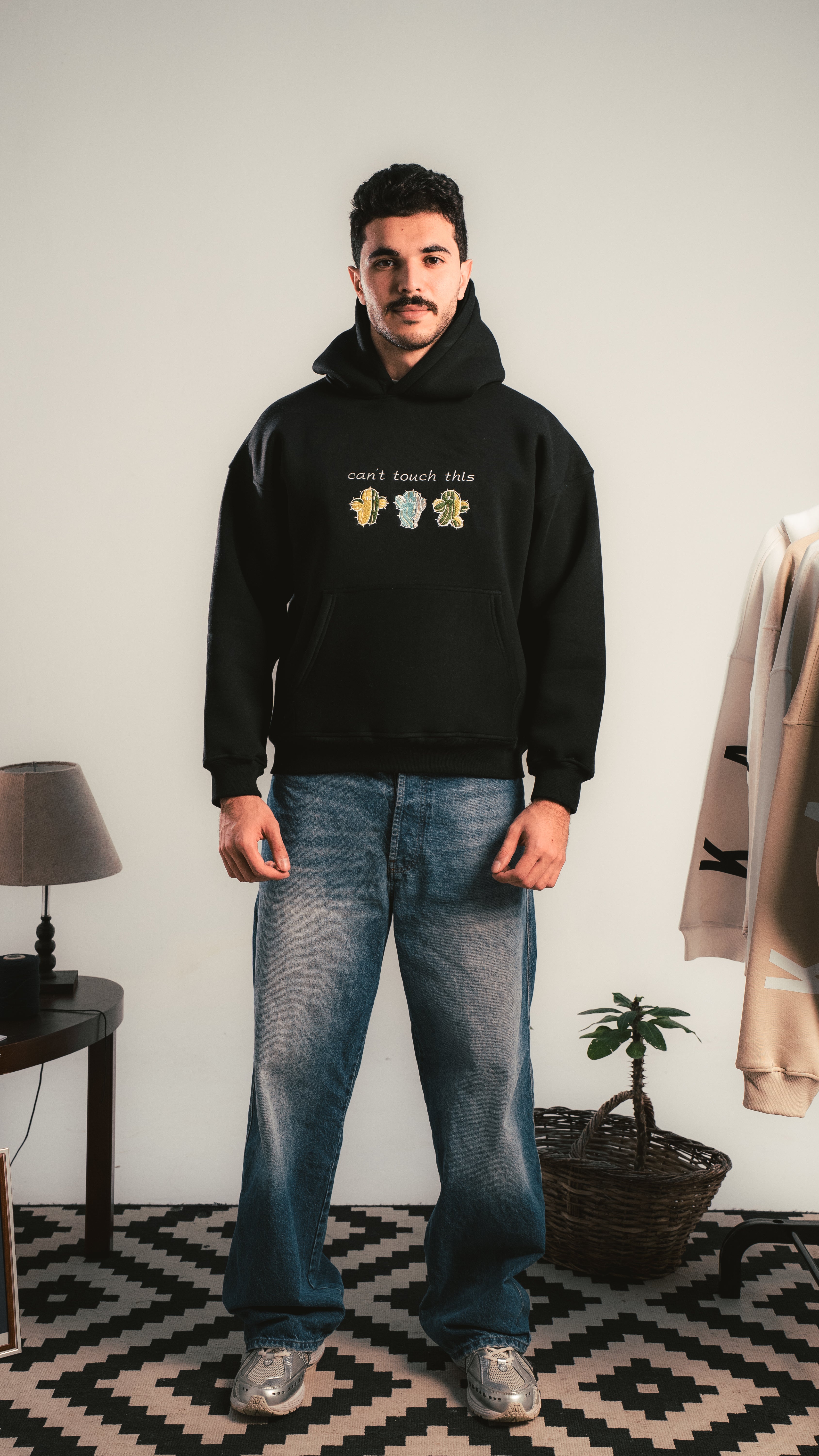 Man wearing a black hoodie with text and graphics, standing in a room with a lamp and coat rack.