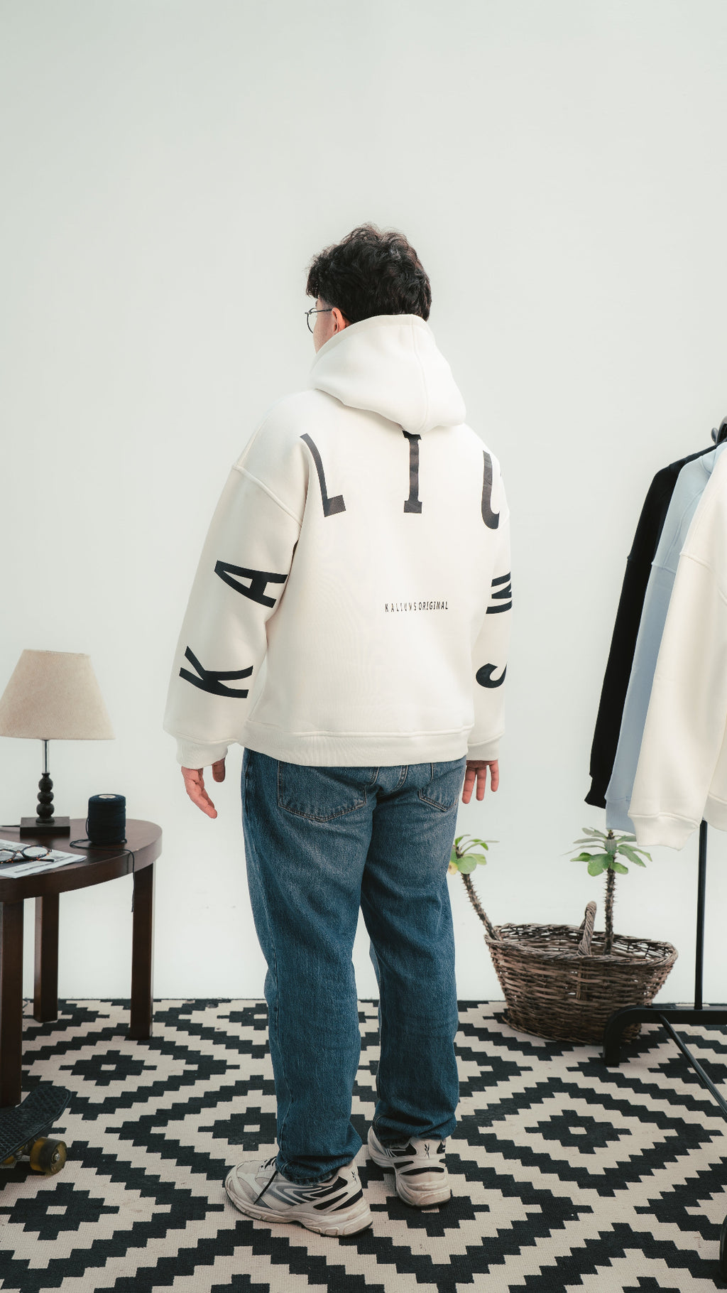 Person wearing a white hoodie with black text in a room with a lamp and plant.