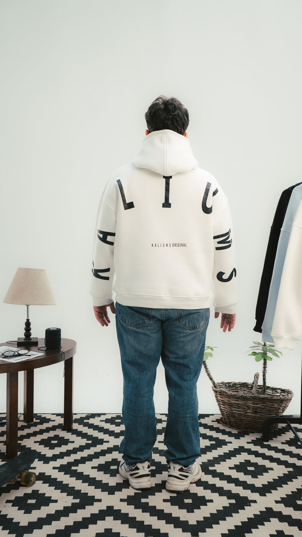 Person wearing a white hoodie with black text standing in a room with a lamp and plant.