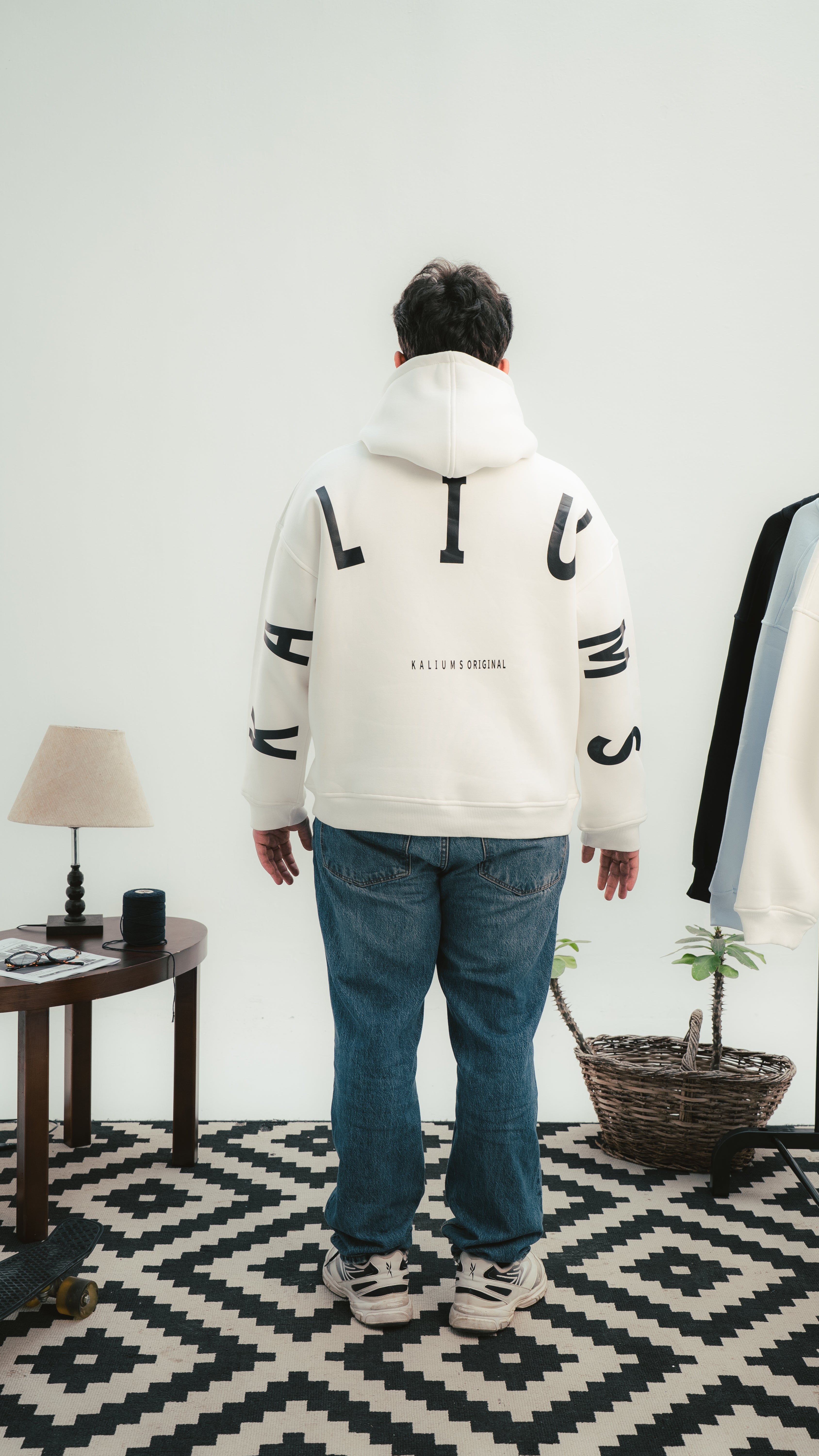 Person wearing a white hoodie with black text standing in a room with a lamp and plant.